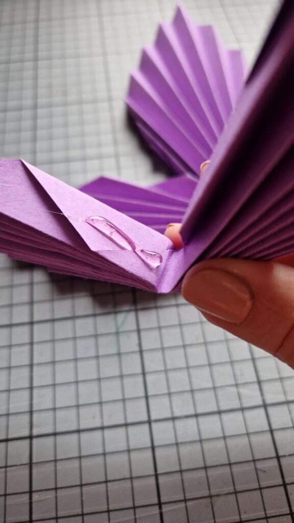 Ein Blatt violettes, zur Ziehharmonika gefaltetes Buntpapier, das im Knick mit Heißkleber zusammengeklebt wird.