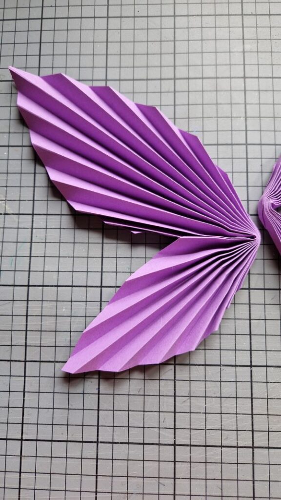 Ein Blatt violettes, zur Ziehharmonika gefaltetes Buntpapier, das im Knick mit Heißkleber zusammengeklebt wurde und so einen halben Schmetterlingsflügel bildet.
