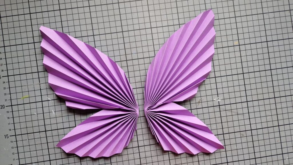 Zwei aus violettem Buntpapier, zur Ziehharmonika gefaltete und in der Mitte zusammengeklebte Schmetterlingsflügel.