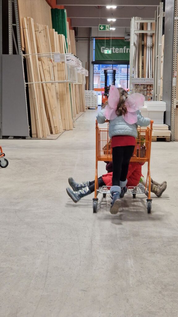 Kinder auf Einkaufswagen im Baumarkt beim Holzzuschnitt.