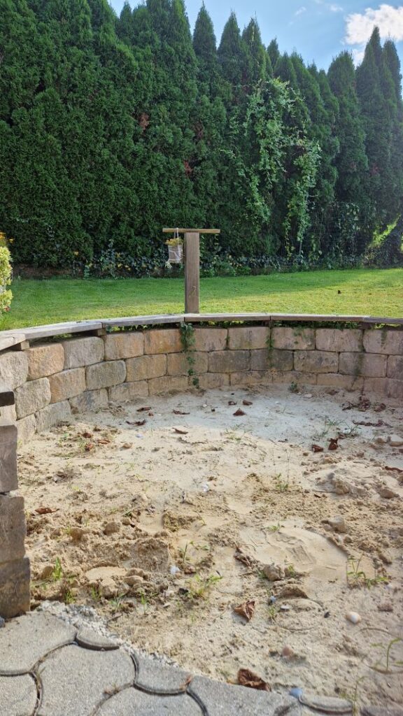 Große, runde Sandkiste die auch als Strandkiste genutzt werden kann und von einer niedrigen Mauer zum Sitzen umrundet wird.